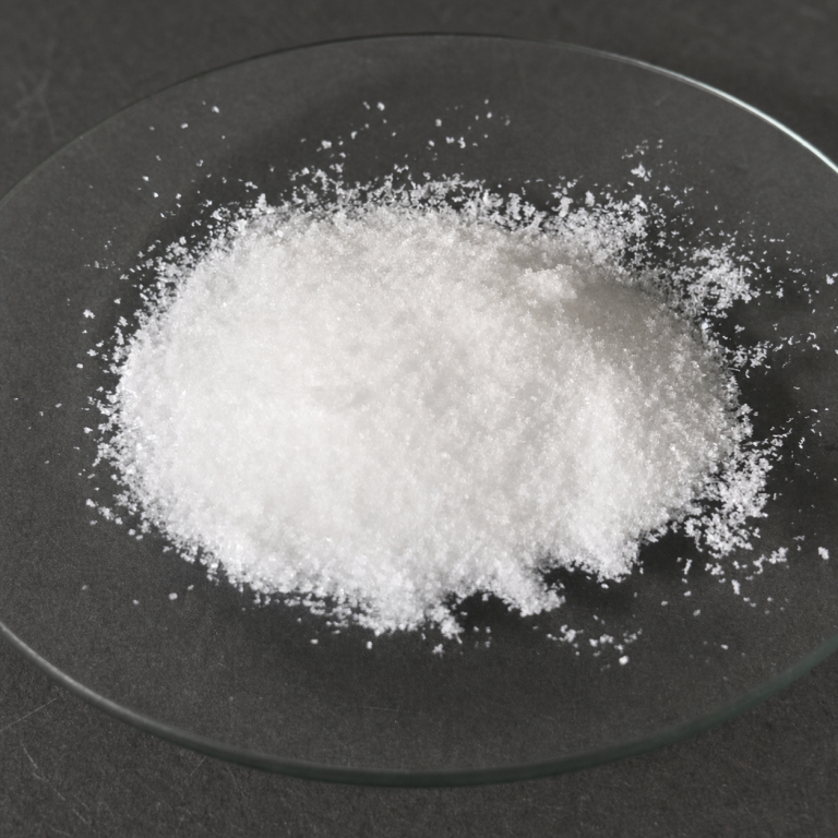 White crystalline benzyltriethylammonium chloride sample displayed on a clear glass dish over a dark charcoal laboratory surface.