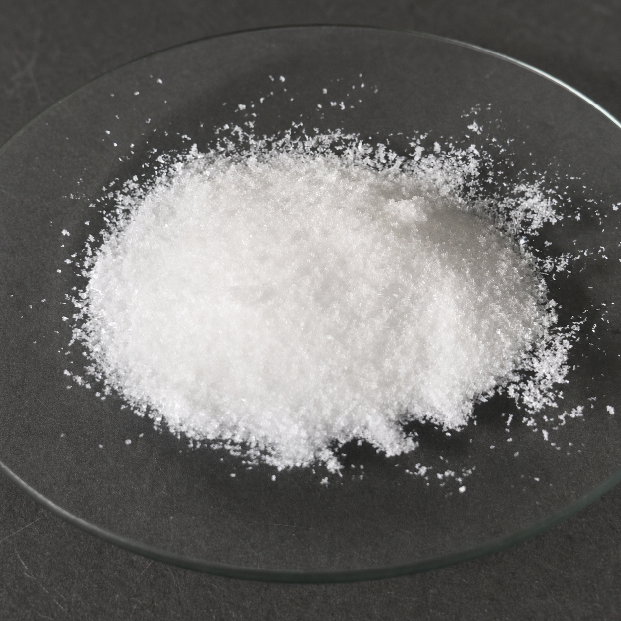 White crystalline benzyltriethylammonium chloride sample displayed on a clear glass dish over a dark charcoal laboratory surface.
