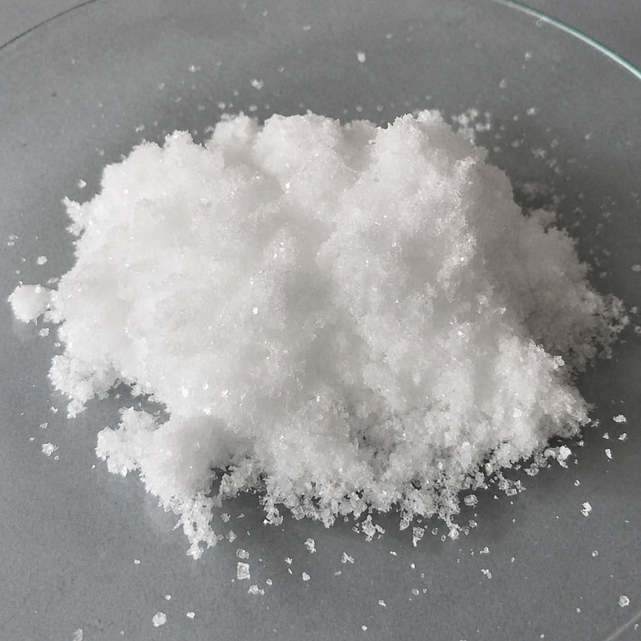 White crystalline tetrabutylammonium bromide sample displayed in a clear glass dish on a cool gray laboratory surface.