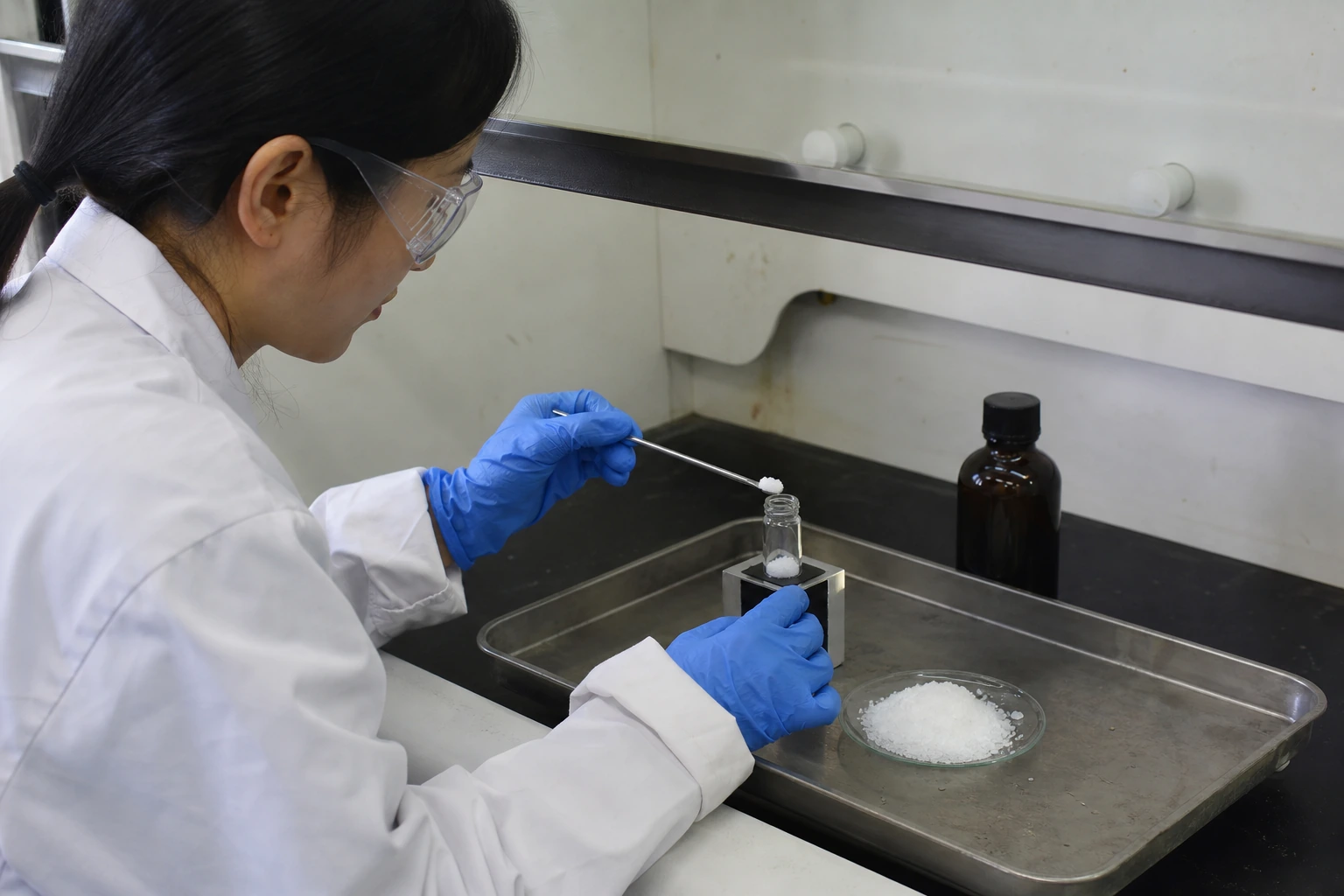 Chinese QC technician transfers white salt-like crystalline material from a glass dish into a supported sample vial inside a fume hood.